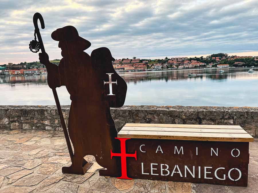 a large metal signboard with a carved out pilgrim for the Camino Lebaniego with the beautiful village of San Vicente de la Barquera in the backdrop